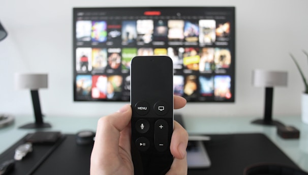 A person holds a remote control in front of a TV displaying a streaming platform interface with various movie and show thumbnails. The room includes a desk with two small lamps on either side of the TV.