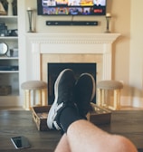 A person is relaxing in a living room with feet up on a wooden table. A TV mounted above a fireplace is displaying a popular streaming platform. Shelves on either side of the fireplace hold decorative items. A smartphone can be seen on the table.