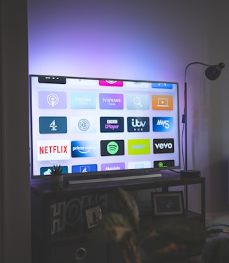 A flat-screen television is displaying a variety of streaming app icons, including Netflix, Prime Video, Spotify, and ITV Hub among others. The TV is placed on a media console with a small potted plant beside it. A tall lamp stands to the right, and a faint, ambient light casts a purple hue on the wall above the TV.