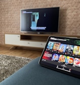 A modern living room features a flat-screen television mounted on a light-colored wooden stand with a patterned wall backdrop. Below the TV, a carpet lies on a wooden floor. In the foreground, a tablet displays a streaming platform interface with various show thumbnails, suggesting a relaxed entertainment setup.